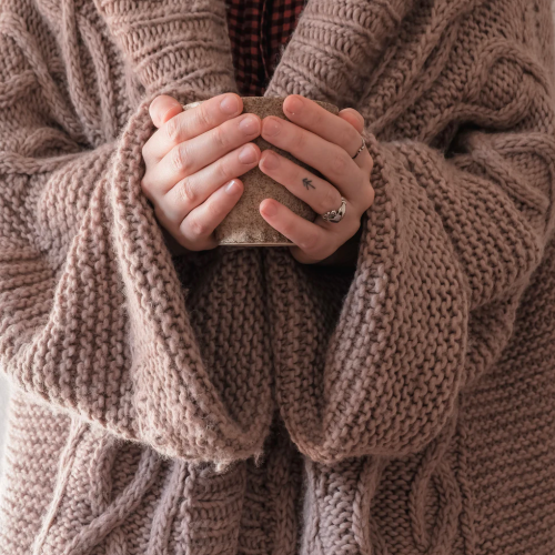 Κοντινό πλάνο ενός ανθρώπου που κρατά ένα ζεστό φλιτζάνι και στα δύο χέρια και φορά ένα μεγάλο πλεκτό πουλόβερ.   Κοντινό πλάνο ενός ανθρώπου που κρατά ένα ζεστό φλιτζάνι και στα δύο χέρια και φορά ένα μεγάλο πλεκτό πουλόβερ.